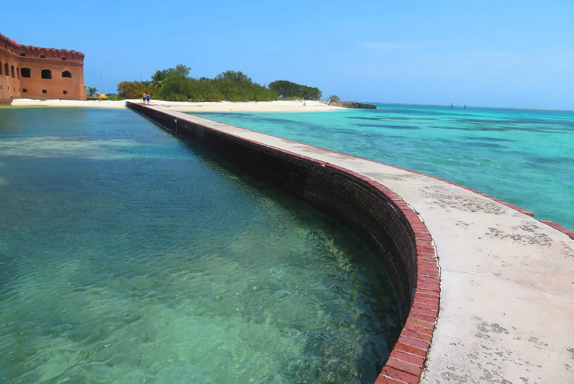 The moat wall at Fort Jefferson on Garden Key, a brick-edged walkway with the moat on one side and clear turquoise sea on the other, the fort's red brick at the far end