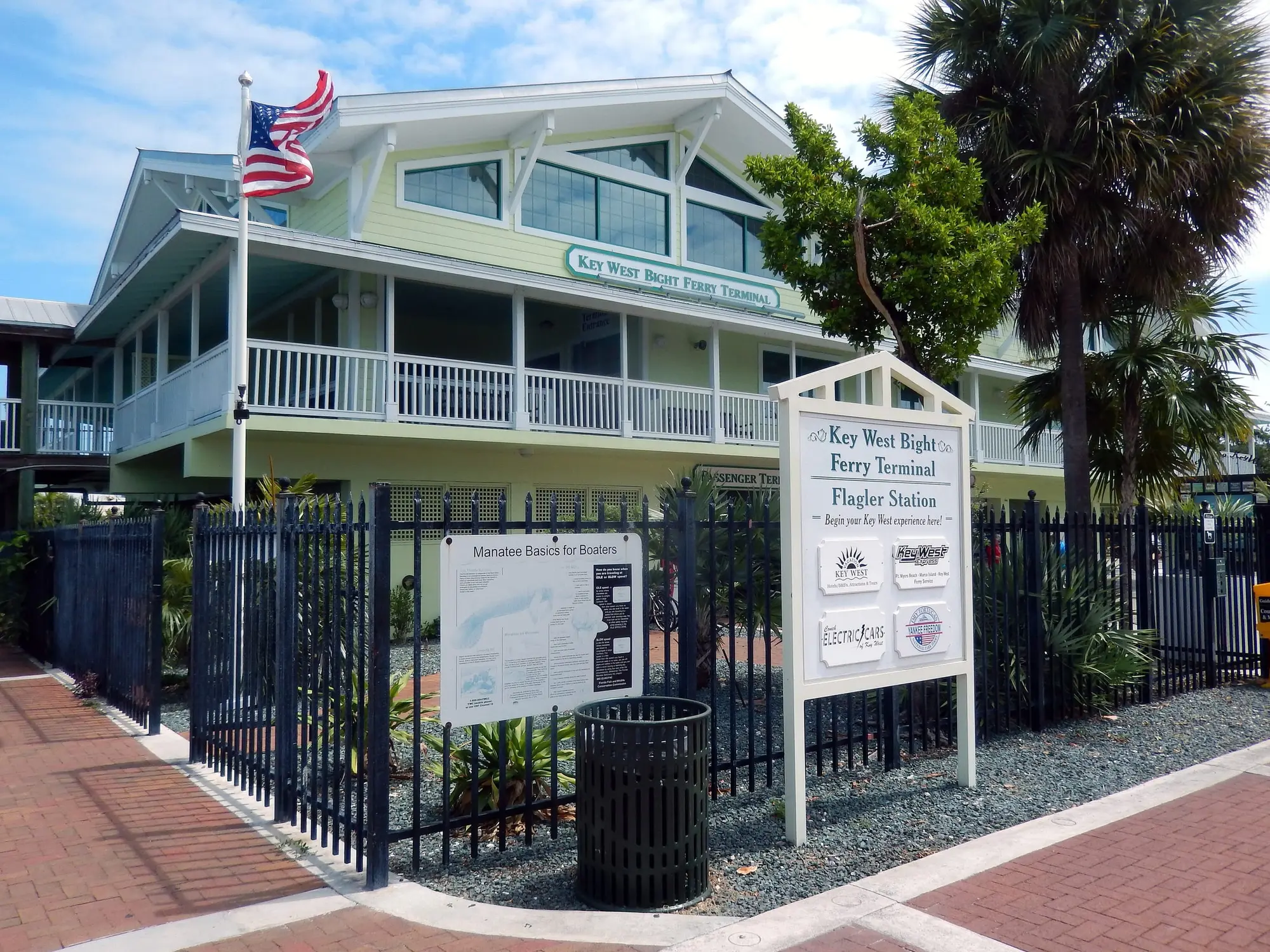 The Key West Bight Ferry Terminal, a pale green two-story building with white porches behind palms, its sign out front at the corner of the brick walk